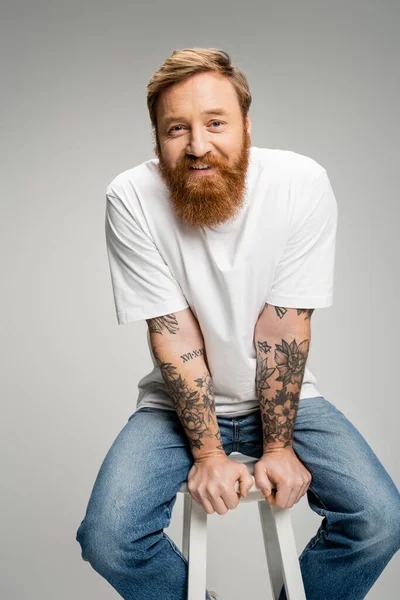Веселий татуйований чоловік у повсякденному одязі сидить на стільці ізольовано на сірому — стокове фото