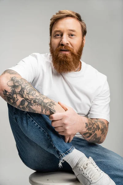 Портрет татуированного мужчины в джинсах и футболке, сидящего на стуле изолированного на сером — стоковое фото