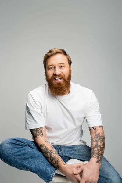 Tattooed man in casual clothes looking at camera while sitting on chair isolated on grey — Stock Photo