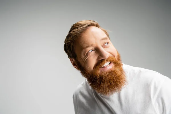 Счастливый бородатый мужчина в футболке смотрит в сторону, изолированный от серых — стоковое фото