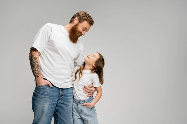 Hombre sonriente abrazando a su hija en camiseta y jeans aislados en gris - foto de stock