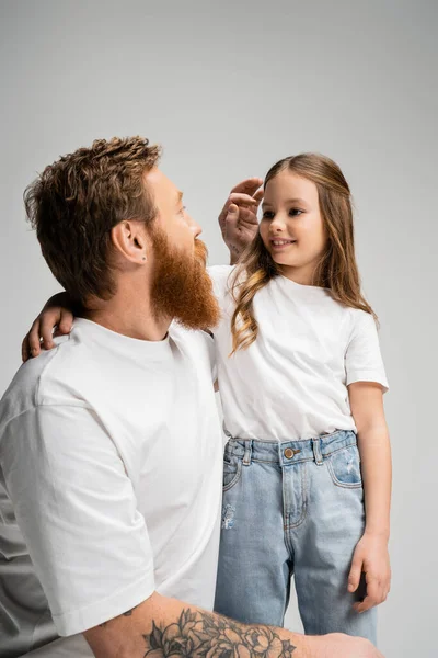 Бородатый отец приспосабливает волосы улыбающейся дочери, изолированной на серой — стоковое фото
