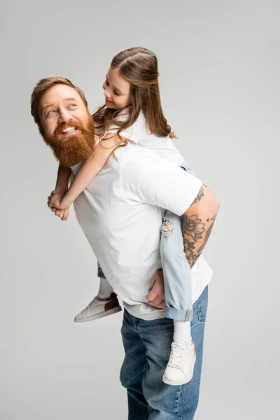 Веселый ребенок, сидящий на татуированном отце с бородой, изолированной на сером — стоковое фото