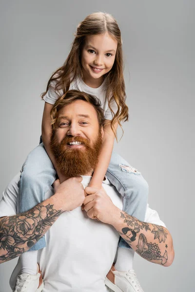 Веселый ребенок, сидящий на плечах бородатого отца, изолированного на сером — стоковое фото