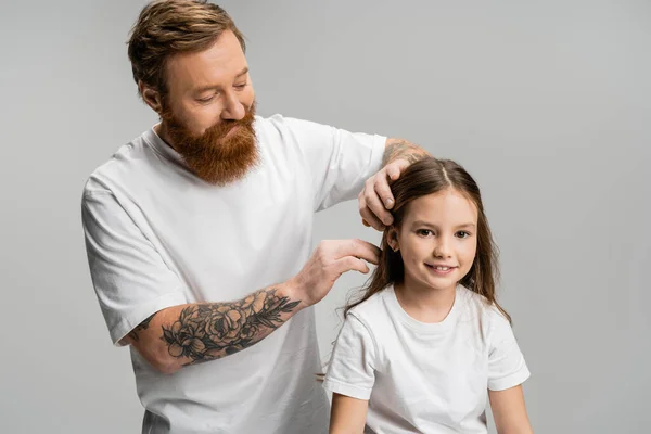 Bearded father adjusting hair of smiling preteen daughter isolated on grey — Stock Photo