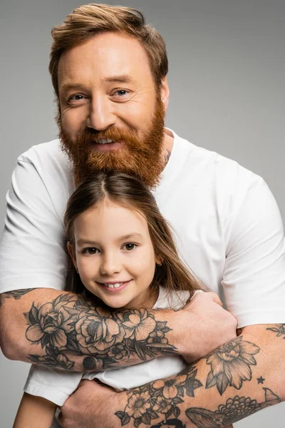 Portrait of tattooed father looking at camera while hugging smiling preteen girl isolated on grey — Stock Photo