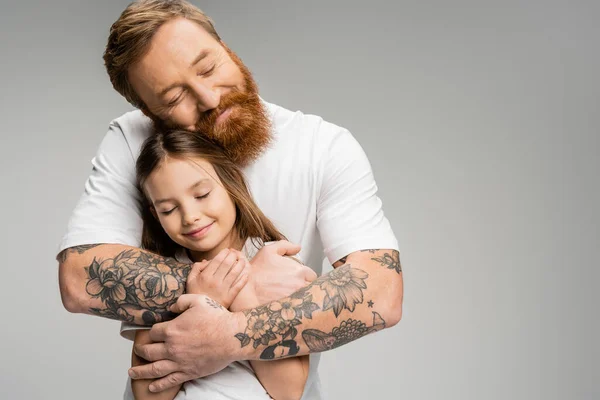 Hombre tatuado en camiseta blanca abrazando a su hija sonriente con los ojos cerrados aislados en gris - foto de stock