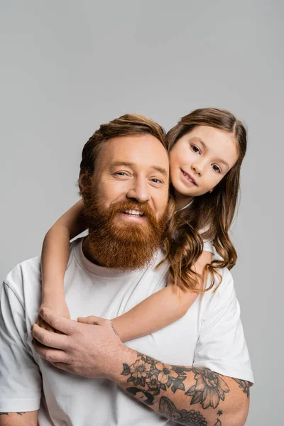 Preteen girl hugging overjoyed tattooed dad and looking at camera isolated on grey — Stock Photo