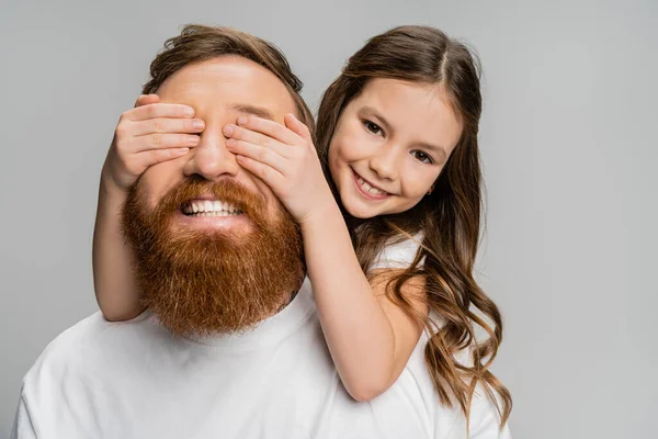 Cheerful preteen girl covering eyes of happy father with beard isolated on grey — Stock Photo