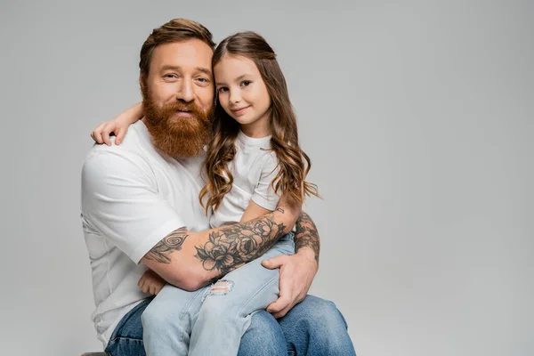 Бородатый мужчина обнимает улыбающуюся дочь в футболке и джинсах, изолированных на серой — стоковое фото
