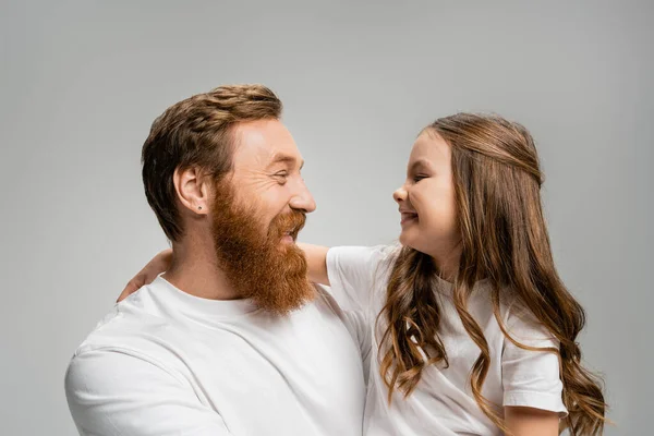 Vista lateral de niña sonriente en camiseta blanca abrazando a papá barbudo aislado en gris - foto de stock