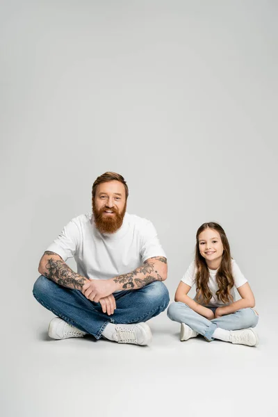 Позитивний татуйований батько і підліток дивляться на камеру, сидячи на сірому фоні — стокове фото