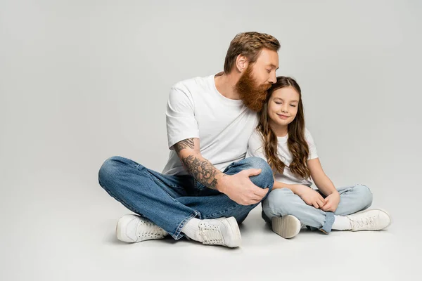 Bärtiger Mann in Jeans und T-Shirt küsst lächelnde Tochter auf grauem Hintergrund — Stockfoto
