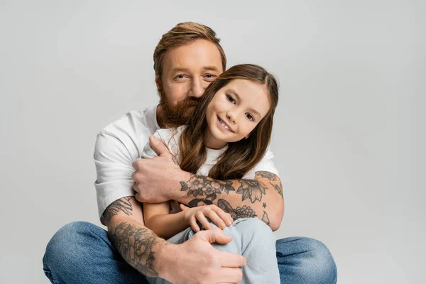 Tattooed man in t-shirt and jeans hugging happy preteen kid isolated on grey — Stock Photo