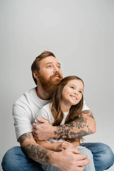 Tatuato uomo abbracciando spensierata figlia e guardando lontano isolato su grigio — Foto stock