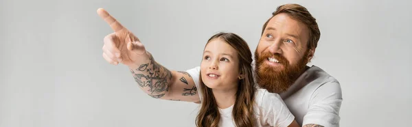 Positive tattooed dad pointing with finger near preteen daughter isolated on grey, banner — Stock Photo