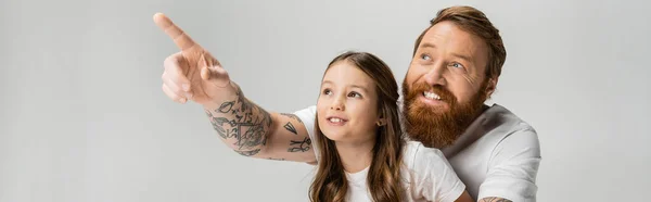 Positivo papá tatuado señalando con el dedo cerca de hija preadolescente aislado en gris, bandera - foto de stock