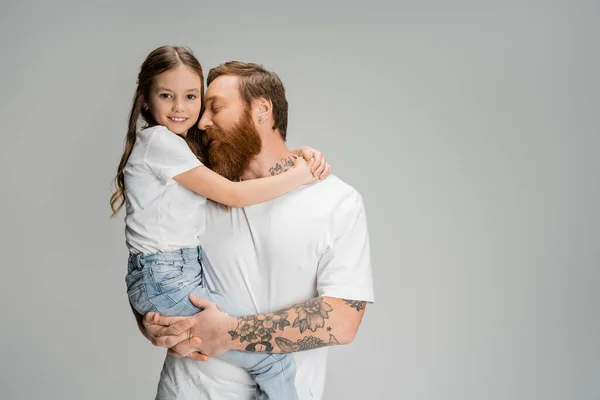 Tattooed father hugging smiling daughter in t-shirt and jeans isolated on grey — Stock Photo