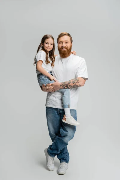 Full length of positive father in t-shirt and jeans holding daughter on grey background — Stock Photo