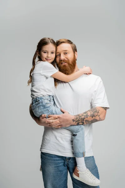 Barbudo y hombre tatuado sosteniendo niño preadolescente y sonriendo a la cámara aislado en gris - foto de stock