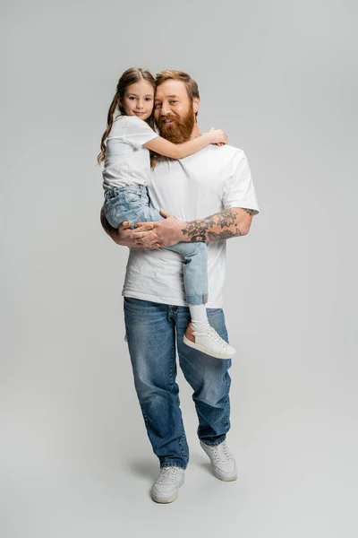 Longitud completa del hombre sonriente en jeans y camiseta que sostiene a la hija preadolescente sobre fondo gris - foto de stock