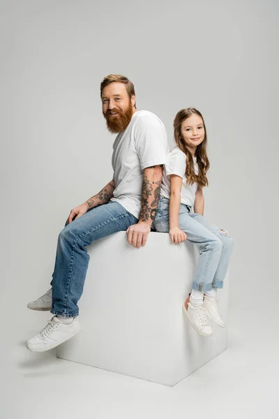 Cheerful tattooed man sitting on cube near daughter on grey background — Stock Photo