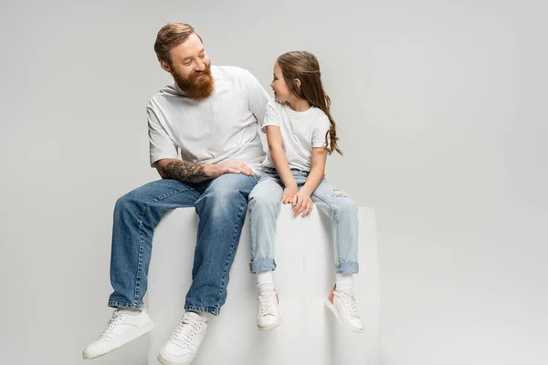 Позитивный татуированный мужчина, смотрящий на дочь в футболке и джинсах, сидящую на кубе, изолированном на сером — стоковое фото