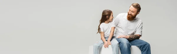 Усміхнена дівчина в джинсах і футболці дивиться на татуйованого батька сидить на кубику ізольовано на сірому, банер — стокове фото