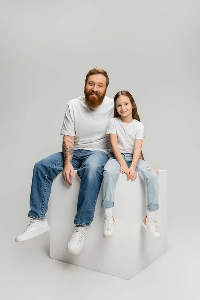 Sorglos sitzen Mann und Tochter auf einem Würfel und schauen in die Kamera auf grauem Hintergrund — Stockfoto
