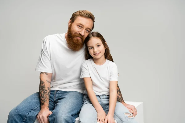 Positivo hombre tatuado sentado cerca de hija en cubo aislado en gris - foto de stock