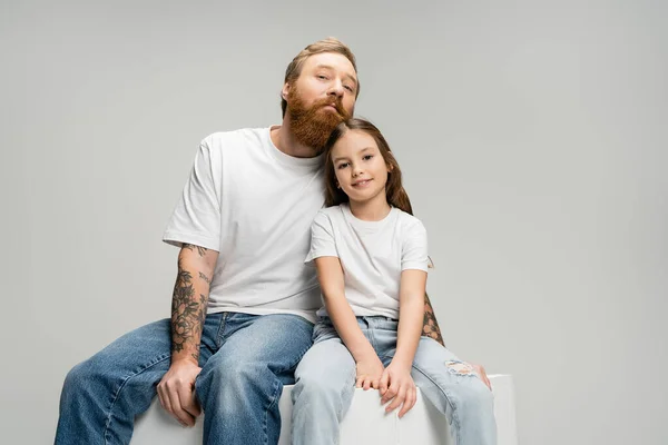 Porträt von Vater und Tochter, die in die Kamera schauen, während sie auf einem Würfel sitzen, der isoliert auf Grau steht — Stockfoto