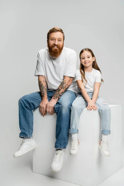 Chica y padre muy alegres sentados en cubo blanco sobre fondo gris - foto de stock