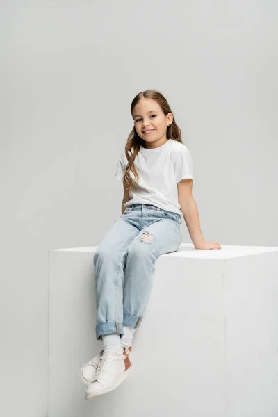 Весела дитина в футболці і джинсах сидить на кубику ізольовано на сірому — стокове фото