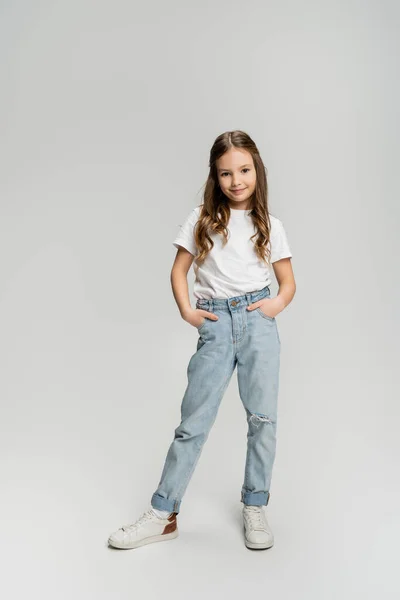 Повна довжина позитивної дитини в джинсах і футболці позує на сірому фоні — стокове фото