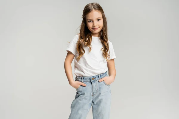 Chica sonriente en camiseta blanca y jeans posando aislados en gris - foto de stock