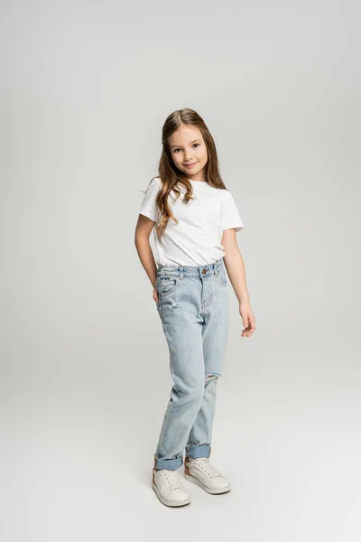 Longitud completa de la niña preadolescente en jeans y camiseta posando y sonriendo sobre fondo gris - foto de stock