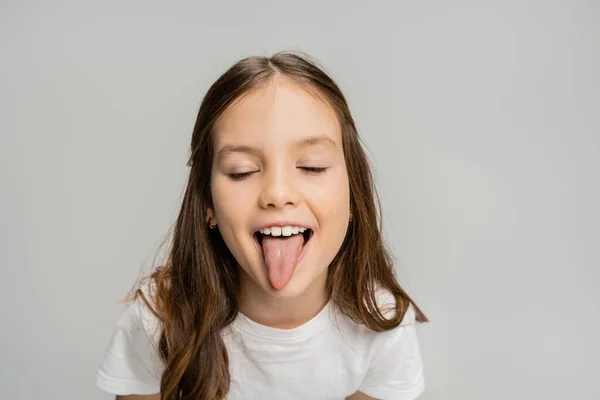 Preteen kid sticking out tongue and closing eyes isolated on grey — Stock Photo