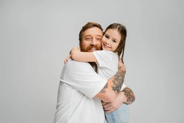 Бородатий чоловік обіймає безтурботну дочку і дивиться на камеру ізольовано на сірому — стокове фото