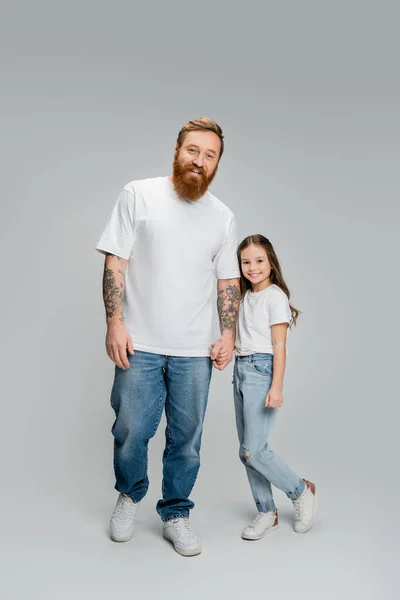 Full length of positive tattooed man holding hand of preteen daughter on grey background — Stock Photo