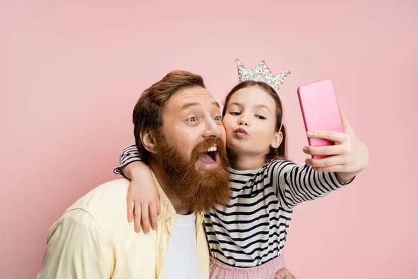 Chica preadolescente con diadema de corona haciendo pucheros labios y tomando selfie con papá barbudo aislado en rosa - foto de stock