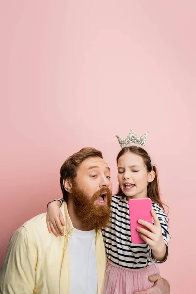 Girl in crown headband taking selfie and hugging dad on pink background — Stock Photo