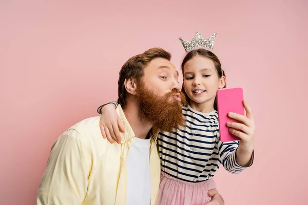 Бородатий чоловік кидає губи, приймаючи селфі з дочкою ізольовано на рожевому — стокове фото