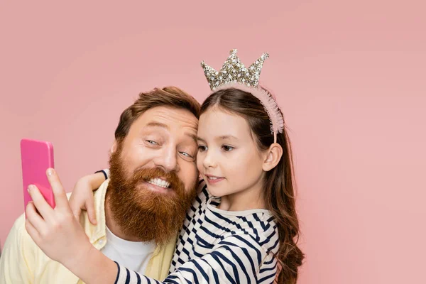 Preteen fille avec couronne bandeau prendre selfie avec gai père isolé sur rose — Photo de stock