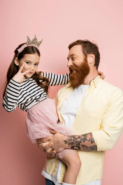 Overjoyed man holding preteen daughter with crown headband showing peace sign on pink background — Stock Photo