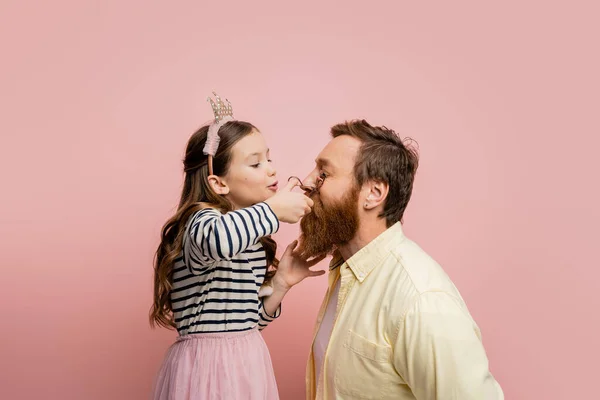 Preteen girl with crown headband curling eyelashes of father isolated on pink — Stock Photo
