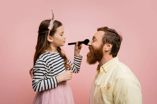 Вид сбоку ребенка в макияжной повязке, держащего макияж рядом с отцом на розовом фоне — стоковое фото
