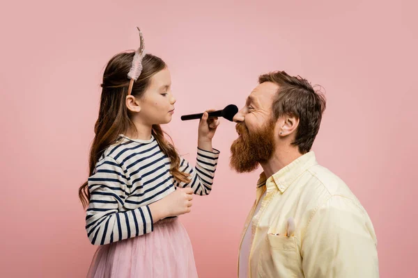 Vue latérale de l'enfant dans le bandeau de la couronne tenant la brosse de maquillage près du père isolé sur rose — Photo de stock