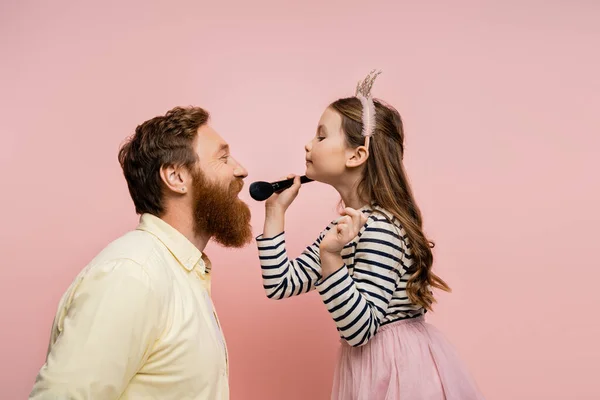 Seitenansicht des Mädchens in Krone Stirnband hält Make-up-Pinsel in der Nähe fröhlichen Vater isoliert auf rosa — Stockfoto