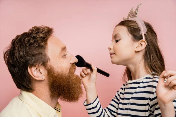 Side view of preteen kid in crown headband applying face powder on bearded father isolated on pink — Stock Photo