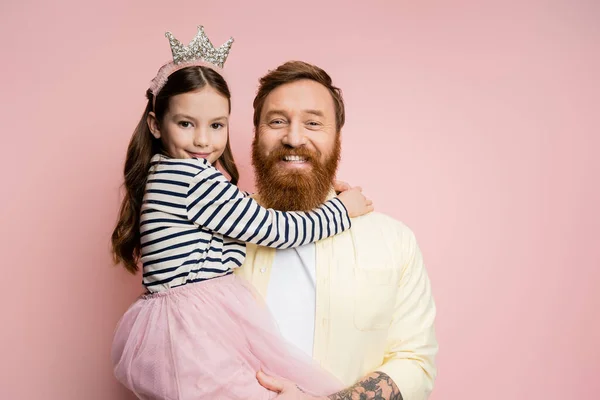 Позитивный и татуированный отец держит дочь с повязкой на голове, изолированной на розовом — стоковое фото